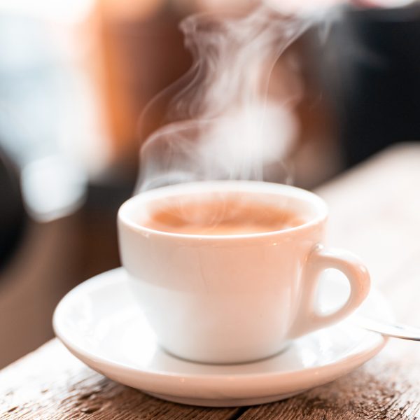 Steam rising from a white cup of hot coffee with a spoon on a saucer over a wooden table in the cafe. Close-up of a refreshing hot cup of a coffee at a cafe.